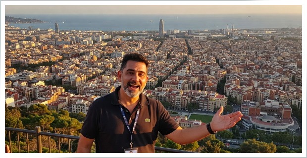 Man with a lanyard smiling and gesturing toward a cityscape from a high viewpoint.
